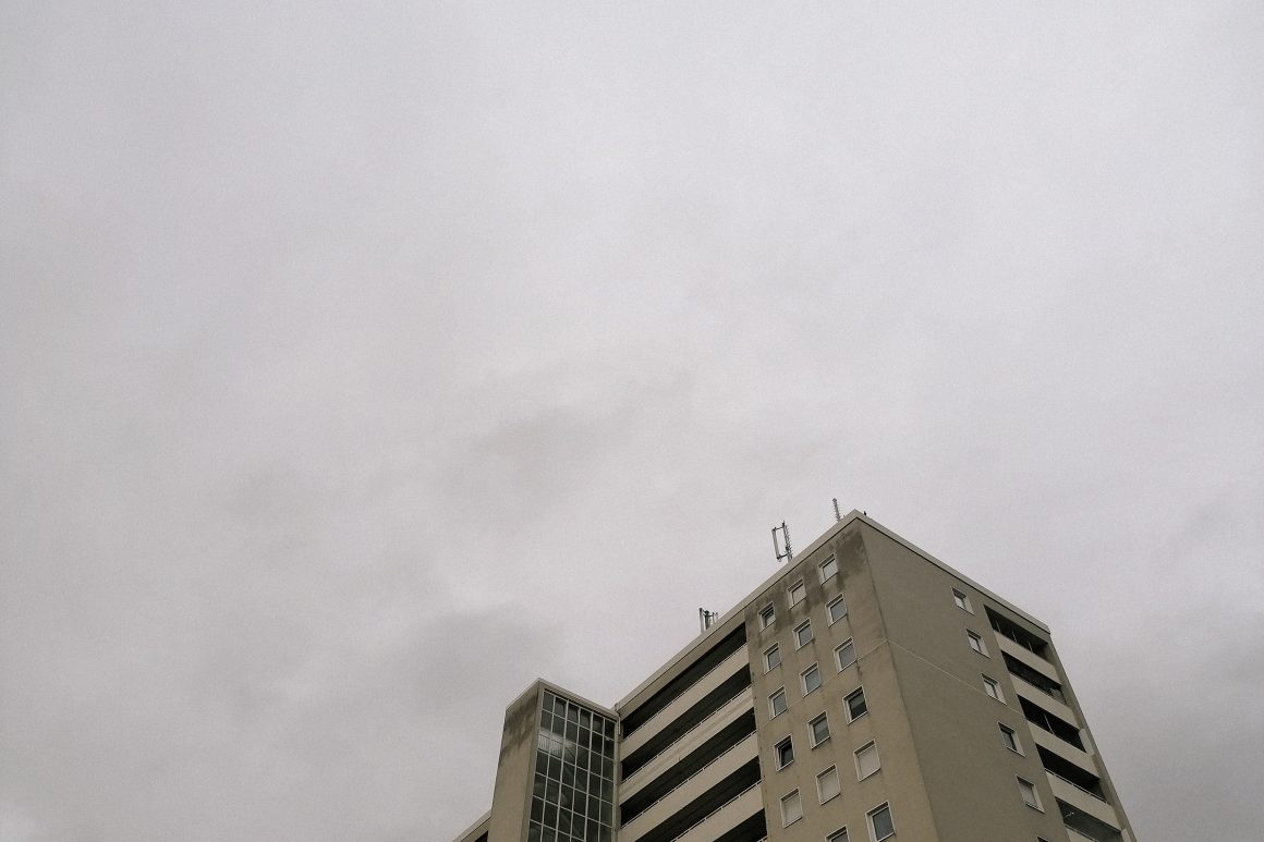 Spitze eines kargen Hochhauses von unten fotografiert, es ragt nur ein wenig in den unteren Teil des Bildes rein. Den größten Teil darüber mein ein HImmel voller Regenwolken aus. 