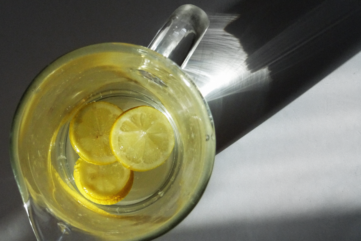 Foto einer Glaskaraffe von oben, darin drei Zitronenscheiben in Wasser; von links strahlt die Sonne hindurch.